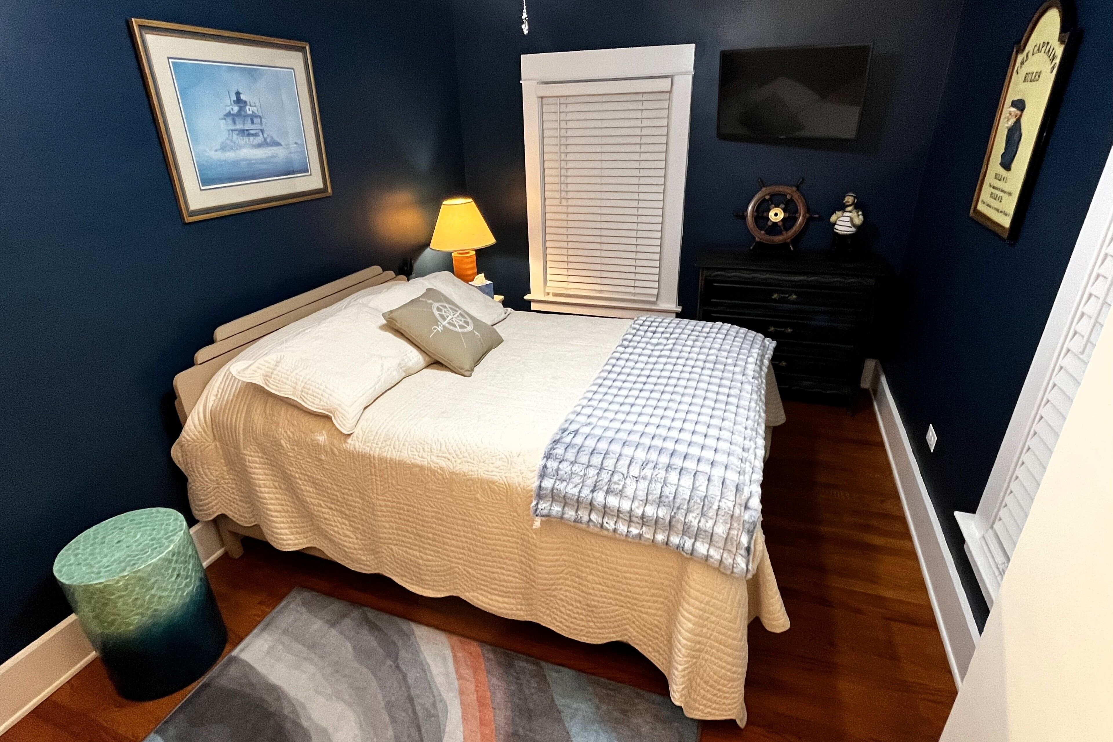 A bedroom with a bed, a nightstand, a lamp, a window with blinds, and a framed picture on the wall.