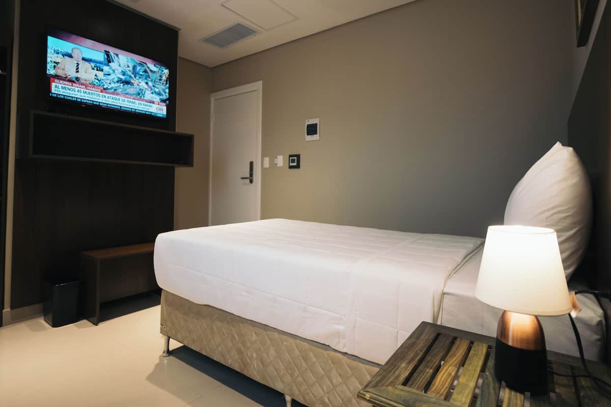 A hotel room with a bed, a bedside lamp, a TV mounted on the wall, and a small wooden table.