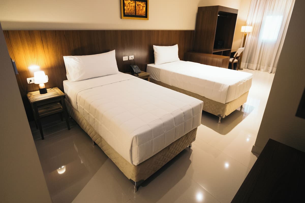 A hotel room with two beds, a bedside table with a lamp, a telephone, a wardrobe, and a painting on the wall.