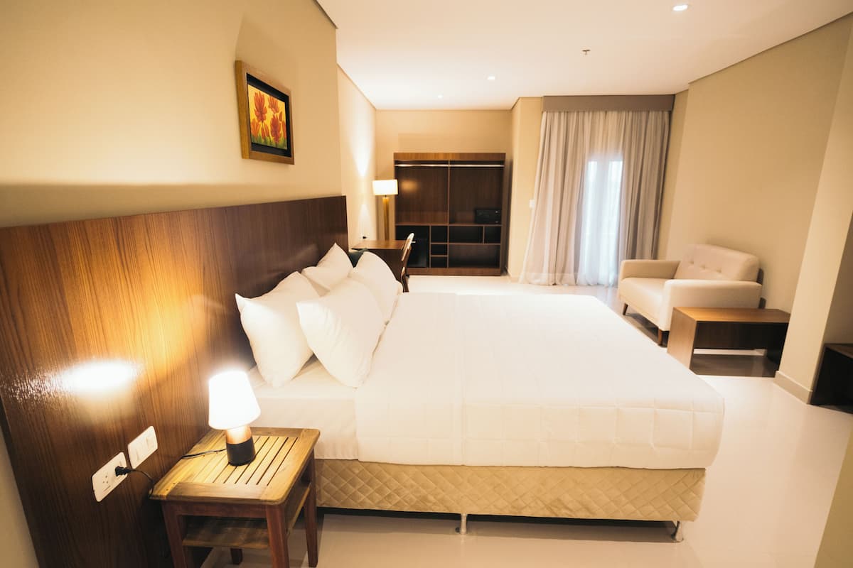 A hotel room with a large bed, a bedside table with a lamp, a chair, a wardrobe, and a painting on the wall.