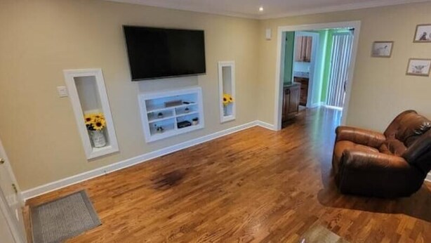 A living room with hardwood flooring, a leather sofa, a mounted TV, and a door leading to a kitchen. - Cardinal Cottage - 2 bedroom cozy home (Muncie)