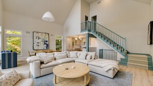 Uma sala de estar ampla com um sofá modular em formato de L, uma mesa de centro redonda e uma escada com corrimão azul-turquesa dando acesso a um andar superior.