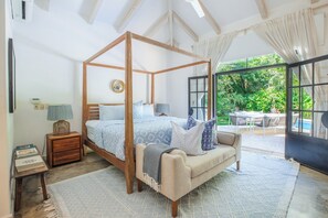 A four-poster bed with a wooden frame and a canopy, a sofa with a throw blanket, a small bedside table with a lamp, and a view of a lush garden through a large window. - Designer 3 bdr Spanish Style Villa with 25m Lap Pool in the Heart of Guiones. (Nosara)