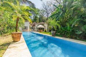 A swimming pool with a tiled edge, surrounded by lush greenery and a building with a tiled roof. - Designer 3 bdr Spanish Style Villa with 25m Lap Pool in the Heart of Guiones. (Nosara)