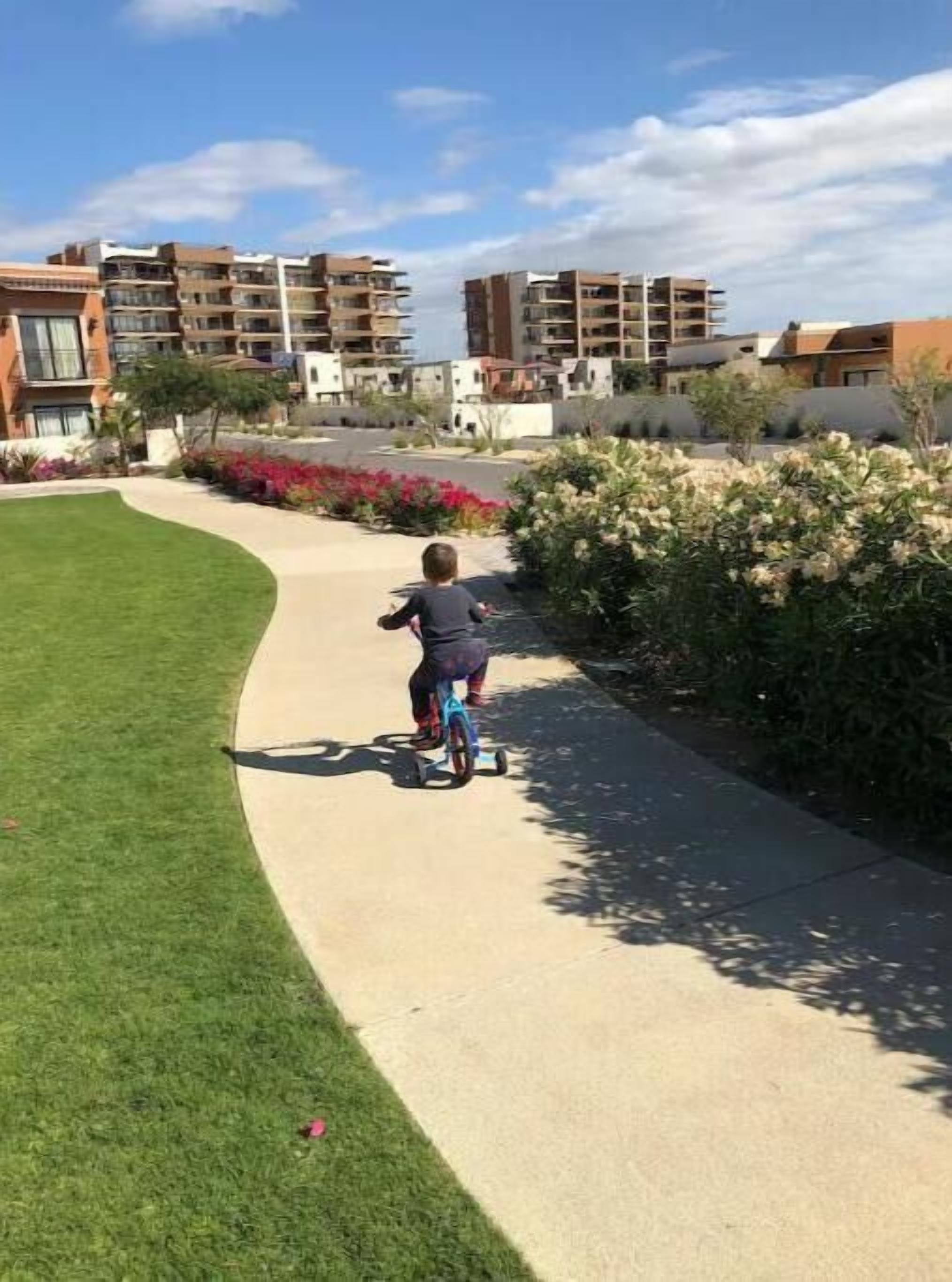 A child riding a scooter on a paved path surrounded by greenery and buildings.