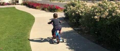 A child riding a scooter on a paved path surrounded by greenery and buildings.