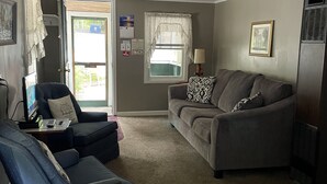 A living room with a gray sofa, a blue chair, a small table with a laptop, and a window with curtains. - Cottage 5 has a very Hamlin lake cottage feel. More private setting.  (Ludington)