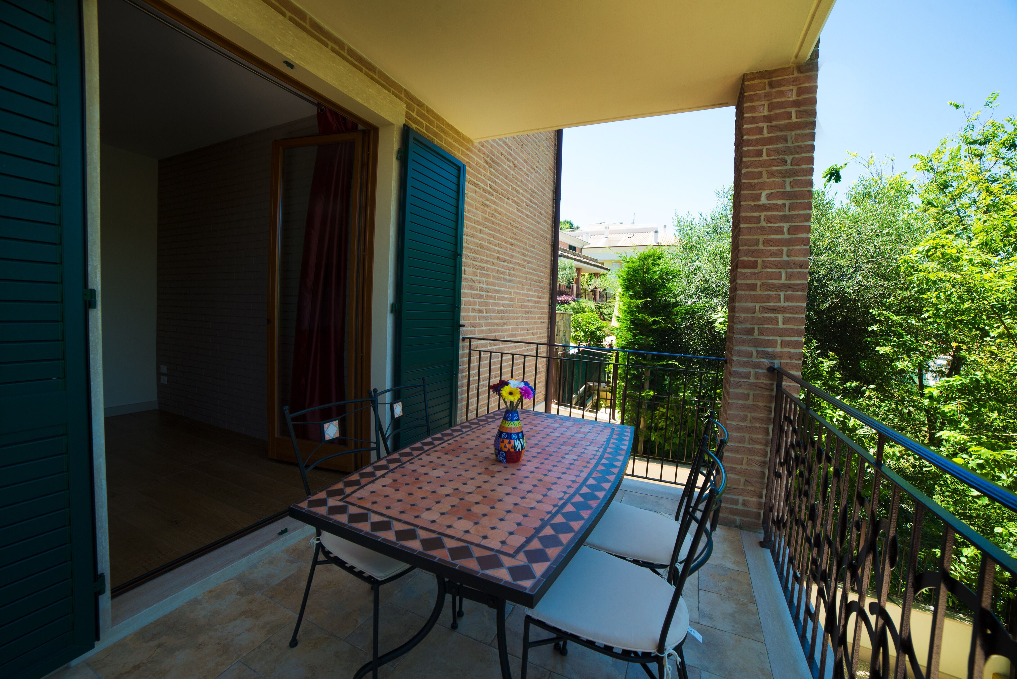 A balcony with a table and chairs, a vase with flowers, and a view of greenery and buildings.