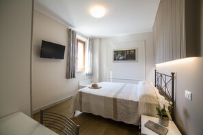 A bedroom with a bed, a bedside table with a plant, a window with curtains, a flat-screen TV on the wall, and a framed picture on the wall.
