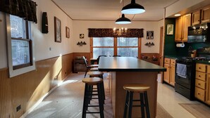 Cozinha com ilha central, banquetas de madeira, mesa de jantar e vista para o exterior através de grandes janelas.