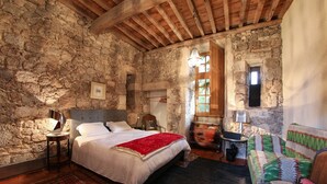 Une pièce rustique avec un mur en pierre, un plafond en bois, un lit avec une literie blanche, un plaid rouge, un tapis à motifs, un canapé vert et blanc, et une petite table avec une lampe.