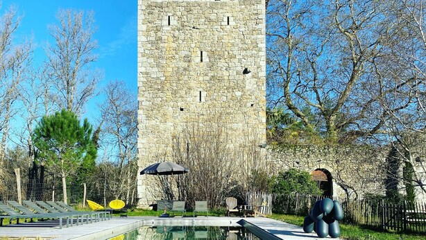 Une tour de pierre avec une piscine devant, entourée d’arbres et dotée d’un espace de détente extérieur.