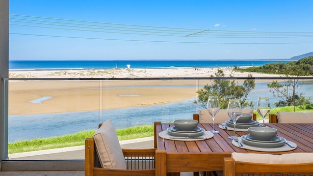 A dining table set with plates, glasses, and cutlery, overlooking a beach and water. - Southcape, Gerroa by the beach (Gerroa)