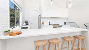 A modern kitchen with a white countertop, bar stools, a stove, and a window with a view of greenery. - Southcape, Gerroa by the beach (Gerroa)