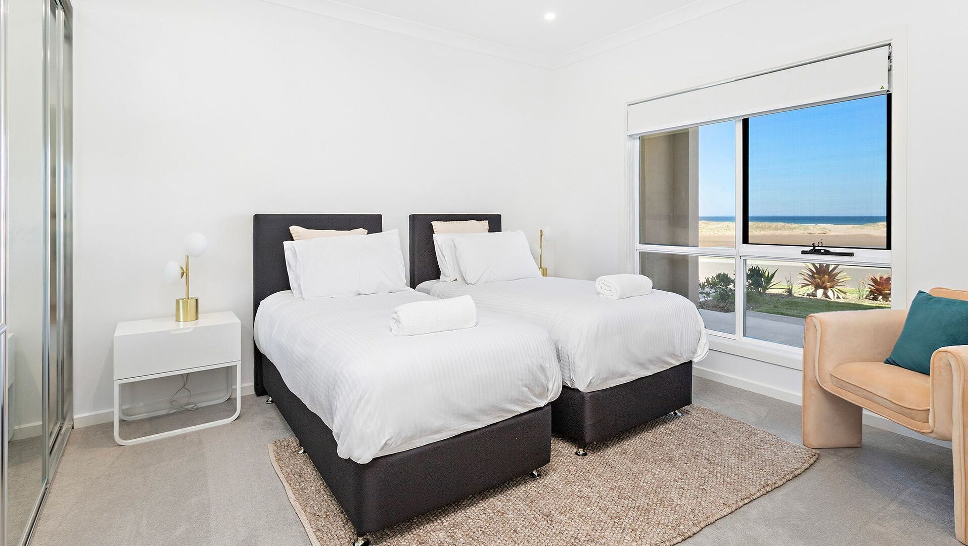A bedroom with two beds, a nightstand, a bedside table, a chair, and a large window with a view of the beach.