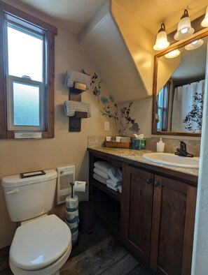 Une salle de bain avec des toilettes, un lavabo et un miroir. Il y a une fenêtre au-dessus des toilettes, et des serviettes sont accrochées à un porte-serviettes.