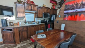 A kitchen with wooden cabinets, a dining table, and a painting of a snowy landscape. - Heigh Ho Tiny Home 30 minutes from Yellowstone  (Island Park)