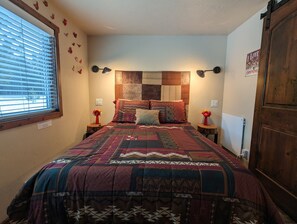 Une chambre à coucher avec un grand lit, deux tables de chevet, une fenêtre avec des stores et une porte en bois.