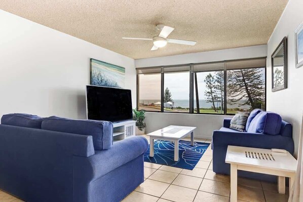 A living room with two blue sofas, a flat-screen TV, a ceiling fan, and a large window with a view of trees and the ocean. - Bellardoo 12 Beach View Mooloolaba (Mooloolaba)