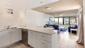 A modern kitchen with white cabinetry, a dishwasher, and a dining area with a blue sofa and a white table. - Bellardoo 12 Beach View Mooloolaba (Mooloolaba)