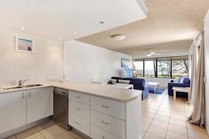 A modern kitchen with white cabinetry, a dishwasher, and a dining area with a blue sofa and a white table. - Bellardoo 12 Beach View Mooloolaba (Mooloolaba)