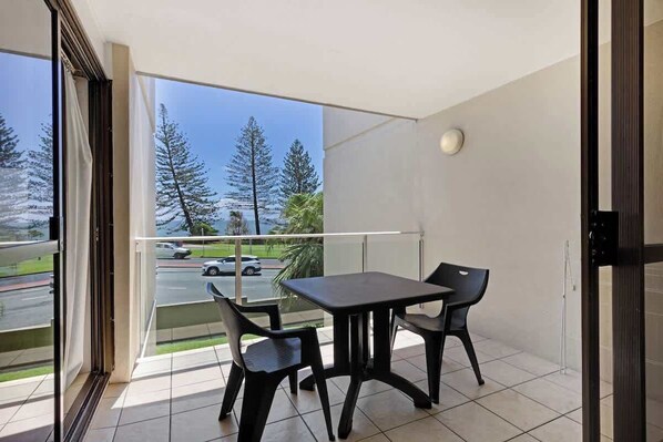 Un balcon avec une table et des chaises, une vue sur les arbres et une route, et une porte donnant sur l’intérieur.