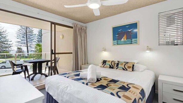 A hotel room with a bed, bedside table, a ceiling fan, a window with blinds, a painting on the wall, and a view of trees and a car outside. - Bellardoo 12 Beach View Mooloolaba (Mooloolaba)
