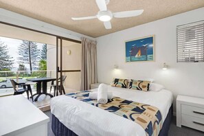 Hotelkamer met een bed, nachtkastje, plafondventilator, raam met jaloezieën, schilderij aan de muur en uitzicht op bomen en een auto buiten.