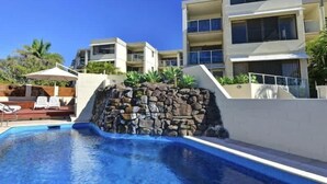 A swimming pool with a stone wall, a lounge area with chairs, and a multi-story building with balconies. - Bellardoo 12 Beach View Mooloolaba (Mooloolaba)
