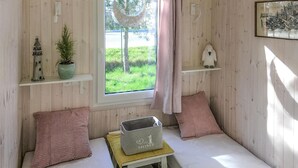 A cozy room with a bed, a small table, a window with a view of greenery, and shelves with decorative items. - Beach front home in Rewal with WiFi (Rewal)