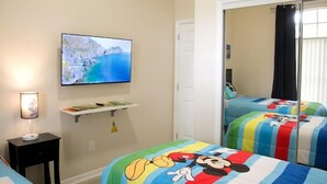 A hotel room with a bed featuring a Mickey Mouse design, a television mounted on the wall, and a window with curtains. - Charming , 2 bedroom , 1 bath  (Kissimmee)
