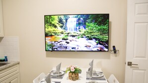 A dining area with a table set for four, a flat-screen TV mounted on the wall displaying a waterfall, and a small vase with flowers on the table. - Charming , 2 bedroom , 1 bath  (Kissimmee)