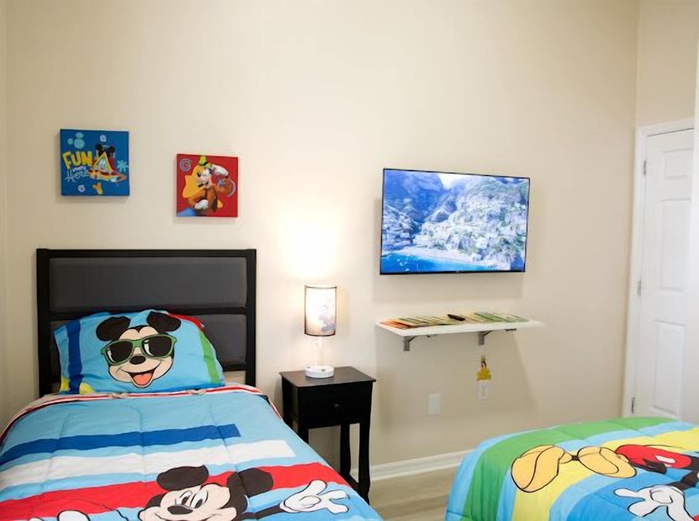 A bedroom with a bed featuring a Mickey Mouse design, a television mounted on the wall, and a shelf with books.