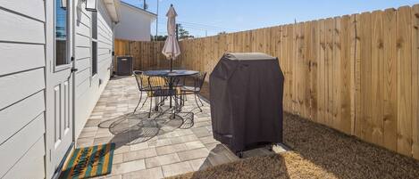 A backyard with a patio, a covered grill, a table with chairs, and a wooden fence.