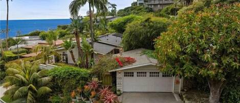A residential area with a house featuring a garage, surrounded by lush greenery and palm trees, with a view of the ocean in the background.