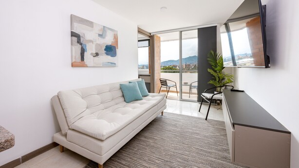 A modern living room with a sofa, a flat-screen TV, a painting, and a view of the city through large windows. - Urban studio in Laureles with nice view near Unicentro Mall (Medellín)