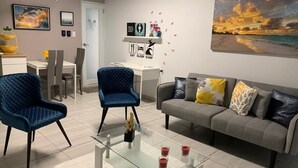 A modern living room with a glass coffee table, two blue armchairs, a grey sofa with patterned cushions, and a painting of a sunset on the wall. - Private Studio, with to much space at the gardens and many amenities like pool. (Toa Alta)
