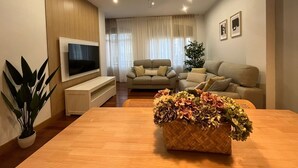 A modern living room with a flat-screen TV, two sofas, a dining table with a centerpiece, and a ceiling fan.