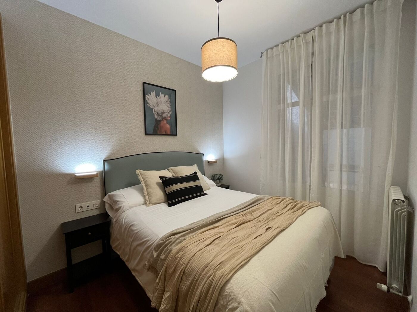 Une chambre à coucher comprenant un lit, des tables de chevet, un luminaire suspendu, une fenêtre avec des rideaux légers et un radiateur.