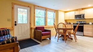 A cozy room with a wooden floor, a dining table with chairs, a sofa, a kitchen area with a microwave and stove, and large windows with a view of trees. - Outdoor Enthusiast's Hideaway and Sauna, Weikert (Millmont)