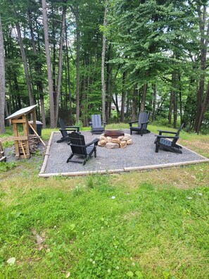 A forested outdoor seating area with four Adirondack chairs, a fire pit, and a wooden structure.