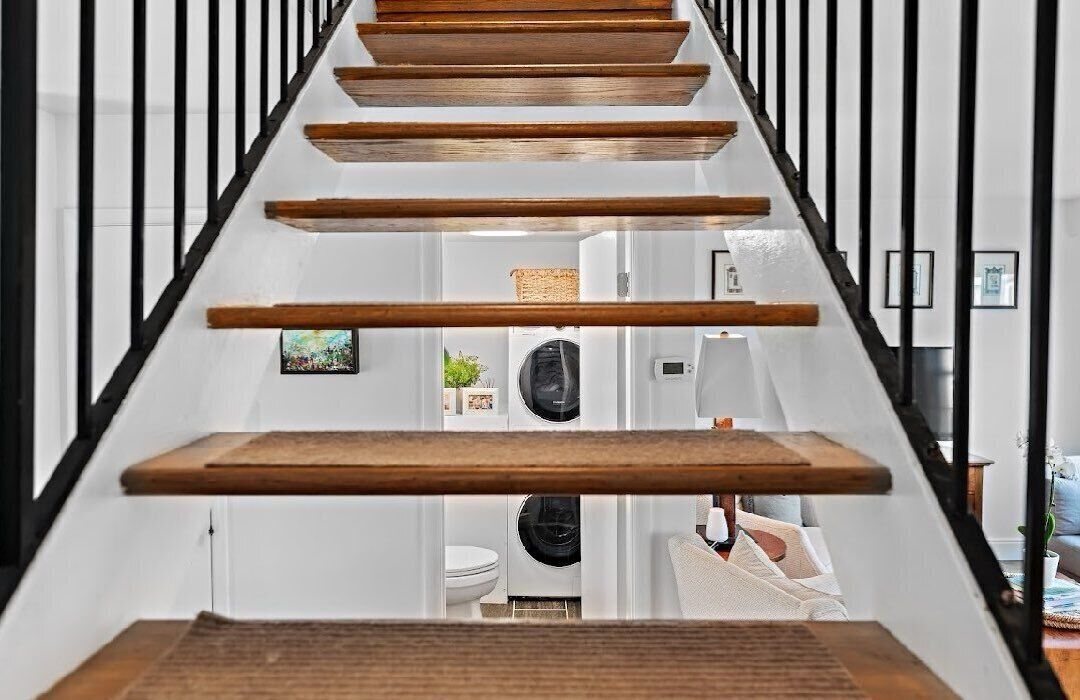 Un escalier moderne avec des marches en bois et des rampes noires, menant à l’intérieur d’une maison où se trouvent une machine à laver et un espace de vie.
