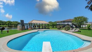 Une piscine agrémentée d’un gazebo à toit de chaume, d’un espace barbecue et offrant une vue sur un immeuble de deux étages.