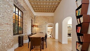 Una habitación con pared de ladrillo, suelo de madera, una mesa con sillas, una estantería y un arco de entrada.