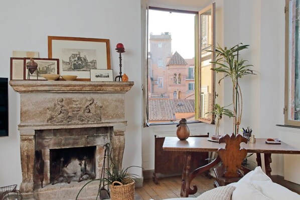 Una habitación con chimenea, una mesa de comedor, sillas, un ventanal con vistas a edificios y una planta en maceta.