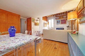 Une pièce avec un plancher en bois, une table à manger recouverte d’une nappe à motifs floraux, un vase bleu et un lit avec une couvre-lit blanche.