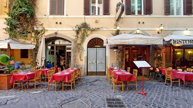 Um café ao ar livre com mesas e cadeiras, uma rua de paralelepípedos e um edifício com porta e janelas.