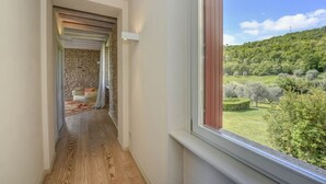 A hallway with a wooden floor leading to a room with a stone wall and a window overlooking a green landscape.
