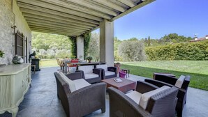An outdoor patio with wicker furniture and a table, surrounded by a garden and a clear blue sky.