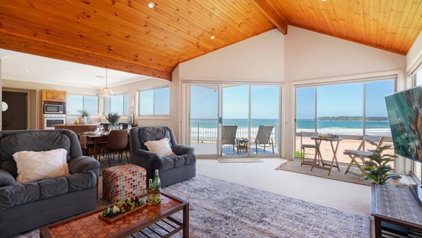 A spacious living room with a large window overlooking the beach, a dining area, and a kitchen. - Dellwood – absolute beachfront on Jones Beach (Kiama Downs)